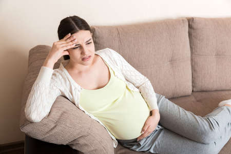 Young pregnant woman with headache sitting on sofa. Pregnancy expectation concept.の写真素材
