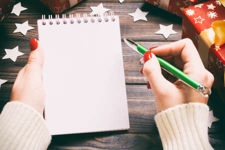 Top view of female hand writing in a notebook on wooden Christmas background. Wish list. New Year concept.の写真素材