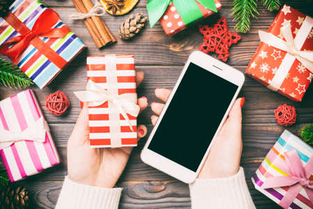 Top view of a woman holding a phone in one hand and a gift in another on wooden background. Fir tree and holiday decorations. Christmas holiday concept. Mockup.の写真素材