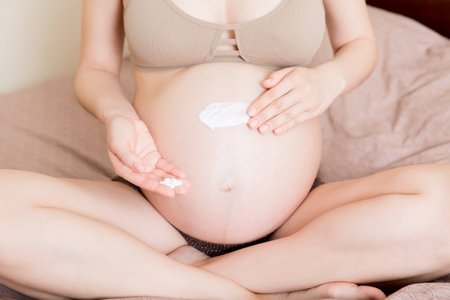 a pregnant girl sits at home on the bed and smears an anti-stretch mark cream on her stomach. Pregnancy, motherhood, preparation and expectation concept.の写真素材