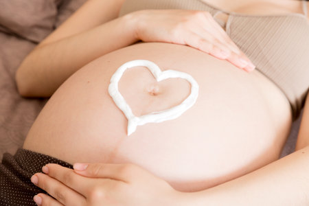 a pregnant girl sits at home on the bed and smears heart an anti-stretch mark cream on her stomach....