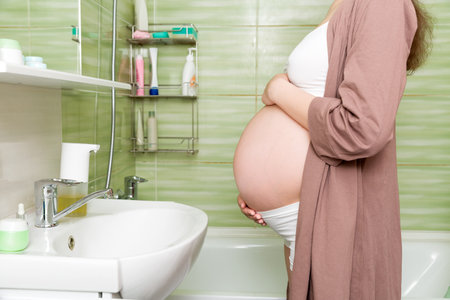 Young pregnant woman dressed in a robe in the toilet at home. Cute adult girl in the third trimester of pregnancy in a home bathroom. Pregnancy, maternity and bodycare concept.の写真素材