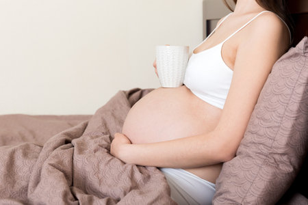 pregnant young woman sitting on bed and drinking coffee at home. maternity concept.の写真素材