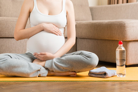 Pregnant young woman with bottle of water while sitting in lotus position on yoga mat. Maintaining water balance at coronavirus time.の写真素材