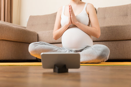 Pregnant female holding smartphone while sitting on exercise yoga mat at home. Fitness or yoga training online at coronavirus time.の写真素材