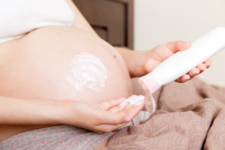 a pregnant girl sits at home on the bed and smears smile an anti-stretch mark cream on her stomach. Pregnancy, motherhood, preparation and expectation concept.の写真素材