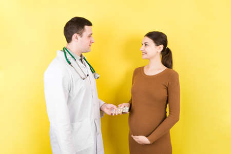Male gynecologist gving pills to pregnant woman during appointment at colored background. Visit To A Doctor Pregnancy Healthcare And People Concept.の写真素材
