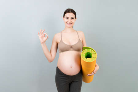 Pregnant Girl Holding Yoga Mat Standing Over Colored Background. Studio Shot, Free Space For Design.