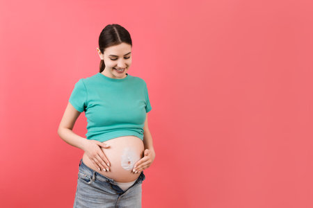 pregnant woman in opened jeans applying moisturizing lotion on her belly to avoid stretch marks at colorful background with copy space. Skin care during pregnancy concept.の写真素材