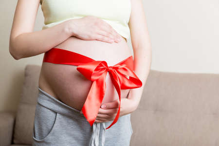 Belly of a pregnant woman tied with a red bow on a home background.