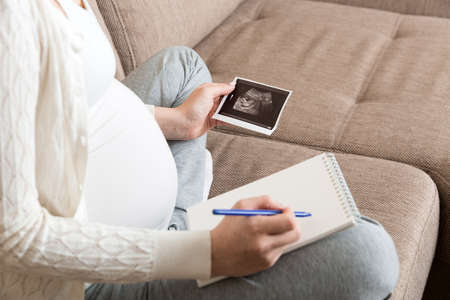 Pregnant Woman sitting on bed and Looking At Ultrasound Scan photo of her Baby. A pregnant female shows her ultrasound report and her unborn baby.の写真素材