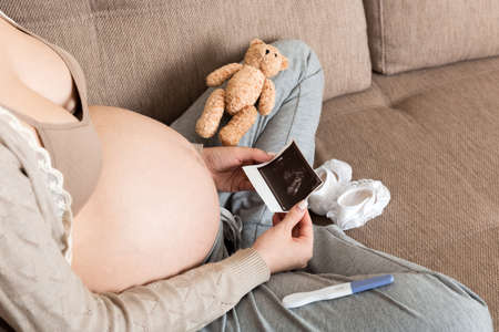 Pregnant Woman sitting on bed and Looking At Ultrasound Scan photo of her Baby. A pregnant female...