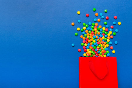 shopping paper gift bag in corner full of assorted traditional candies falling out on colored background with copy space. Happy Holidays sale concept.の写真素材