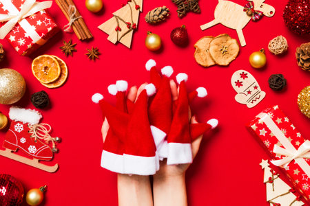 Top view of female hands holding a stack of santa hats on red background. New year decorations. Christmas holiday concept.