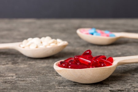 Vitamin capsules in a spoon on a colored background. Pills served as a healthy meal. Red soft gel vitamin supplement capsules on spoon.の写真素材