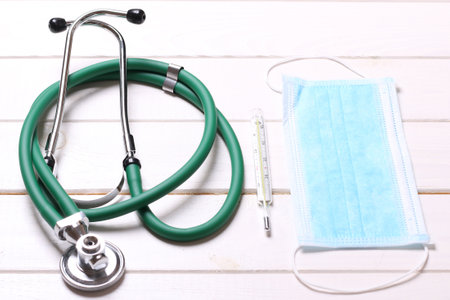 Top view of stethoscope, protective mask and mercury thermometer on white wooden background. Medical...