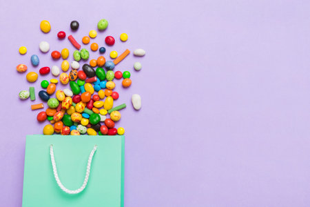 shopping paper gift bag in corner full of assorted traditional candies falling out on colored background with copy space. Happy Holidays sale concept.