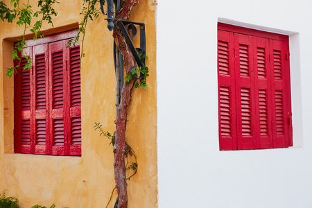 Two red wooden windows one at white wall the other at yellow wall with tree and plantsの写真素材