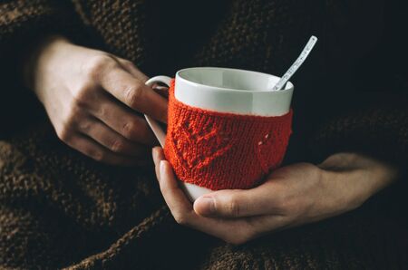 Red Knitted woolen cup with heart pattern in female hands. Hands holding a cozy Mug in red knitted mitten with hot cocoa, tea or coffee. Valentines day conceptの写真素材