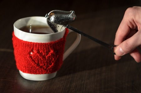 Mug in red knitted mitten with heart pattern on wood tabl and Hand with spoon for tea leaveson. Tea brewing conceptの写真素材