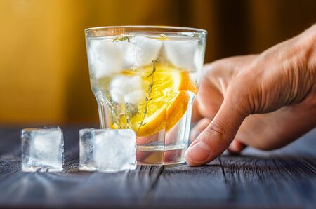 Alcoholic drink with lemon and ice in a glass on a old dark wooden table. Close up view of cold drink with human handの写真素材
