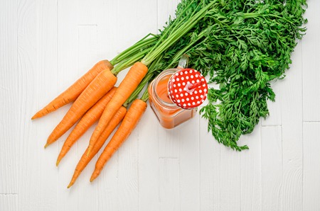 Healthy carrot smoothie in a jar with a red lid in a white polka dots and an orange straw and carrots on white wooden table top view.の写真素材