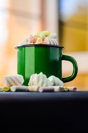 The green enameled mug is filled with a multicolored marshmallow on a black wooden table with blurred interior background close-upの写真素材