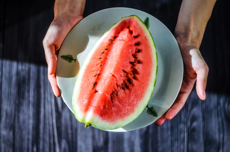 Slice of watermelon on white plate in woman hands ready for eating, top view with blurry black wood table backgroundの写真素材