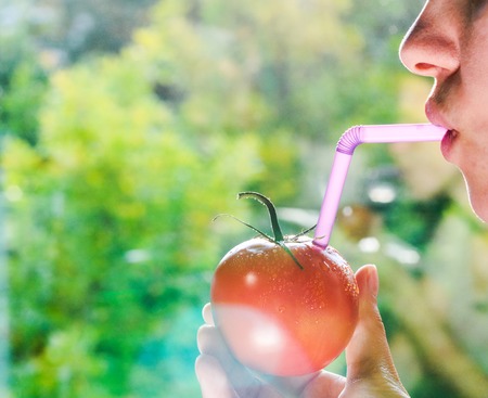 Girl Drink Juice From Tomato Through a Straw on Blurry Green Background. Concept of Fresh Juice Straight from Vegetables. Tomato as a Vial of Juiceの写真素材