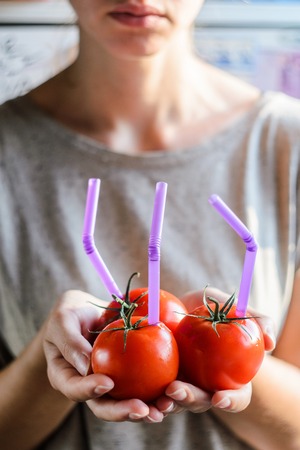 Three Ripe Red Tomatoes with Straws in Woman Hands on Bright Background. Fresh Juice Healthy Food Concept. Tomato as a Vessel with Fresh Juiceの写真素材