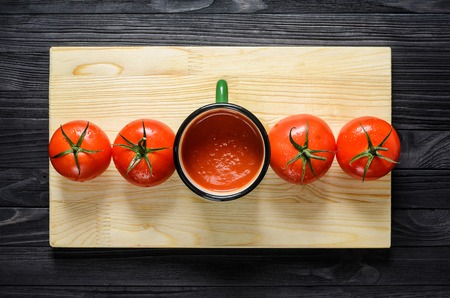 Tomato Juice in Green Enamel Mug on a Wooden Cutting Board on Black Wooden Table with Four Fresh Tomatoes Top View.の写真素材