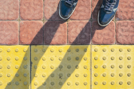 View from the First Person to his Feet, Standing in Front of the Sidewalk with Tactile Tiles to Navigate Blind People. Blue Sneakers on Yellow Tactile Paving Slabs.の写真素材