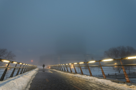 Silhouette of a man passing through a pedestrian bridge in the dark in a strong fog. Handrails with integrated lamps illuminate the way for himの写真素材