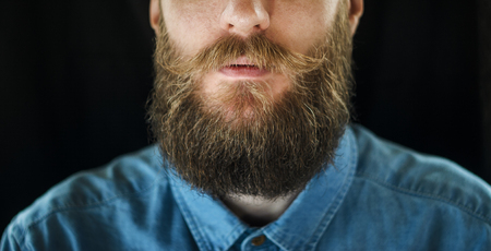 Attributes of the Appearance of a Hipster Male: Mustache and Beard Close Up. Portrait of Bearded Man in a Blue Denim Shirt on Black Backgroundの写真素材