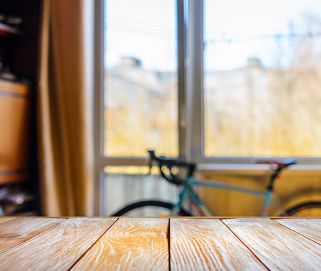 Empty Wooden Table in Front of Blurred Background of Living Room with a Blue Bicycle Near the Window. Mock Up for Display of Product. Blank for Your Layoutの写真素材