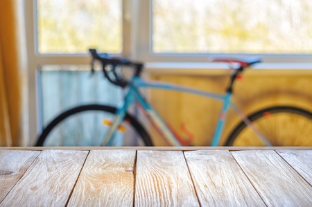 Empty Wooden Table in Front of Blurred Background of Living Room with a Blue Bicycle Near the Window. Mock Up for Display of Product. Blank for Your Layoutの写真素材