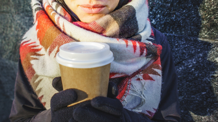 Young beautiful girl in gloves and in a large cozy scarf holding a paper cup of takeaway coffee. Coffee to go in winter conceptの写真素材