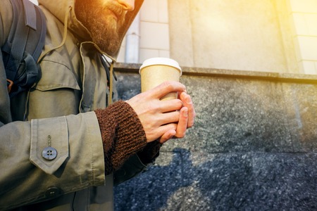 Paper cup of hot coffee in the hand of a bearded man in a jacket and a brown sweater. Warming drink in cold winter weather. Take away coffee concept. Coffee cup layoutの写真素材
