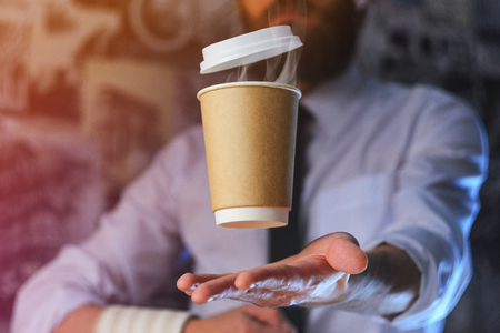 Barista in a white shirt with a tie holds floating in the air a paper cup with a hot drink on his outstretched arms. Barista wizard concept. Background for advertising and placing logo on your drinkの写真素材