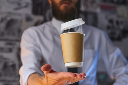 Levitating in the air paper cup with hot coffee. Barista, a bearded young man in a white shirt with a tie, creates miracles - advertises his drink, causing it to soar. Logoplacement conceptの写真素材