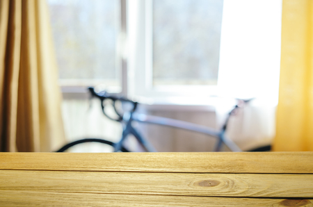 Product Placement Background. Wooden Table Against Blurry Interior of Cyclist's Living Room with Large Window and Blue Sport Bicycleの写真素材