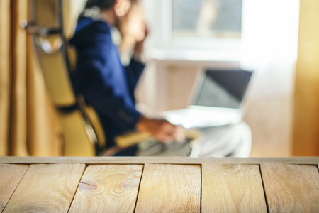 Wooden Table Top Against Blurry Interior Background. A Bearded Man in a Blue Jacket Works for Laptop. Freelance Work Concept. Blank for Your Layoutの写真素材