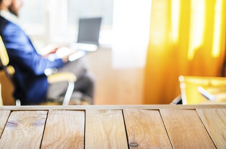 Wooden Table Top Against Blurry Interior Background. A Bearded Man in a Blue Jacket Works for Laptop. Freelance Work Concept. Blank for Your Layoutの写真素材