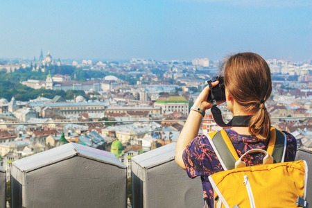 Young Girl Tourist Photographer with a Yellow Backpack Takes Pictures of Sights. Travel Concept. Panorama of the Old Townの写真素材