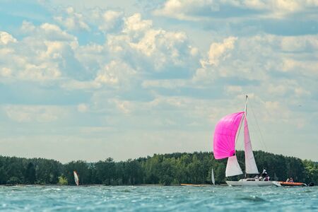 A small sailing yacht with a pink spinnaker and white sails goes to the bay on a sleepy day against...