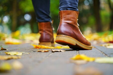 legs of a woman in black pants and brown boots walking in a park along the sidewalk strewn with...