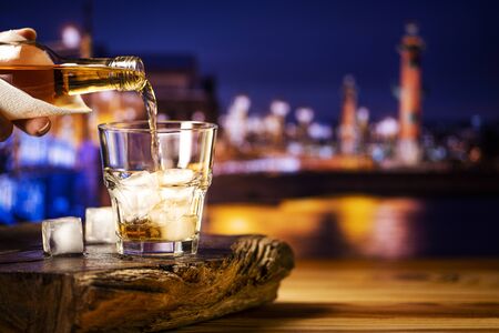 Pouring whiskey into a glass with ice against the background of the night city. A glass of alcoholic beverage on a wooden table.の写真素材