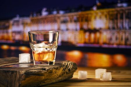A glass of alcoholic beverage on a wooden table against the background of the night city. Whiskey cocktail with iceの写真素材