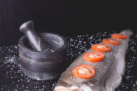 White fish with tomatoes and marble mortar on a black wooden background. Process of cookingの写真素材