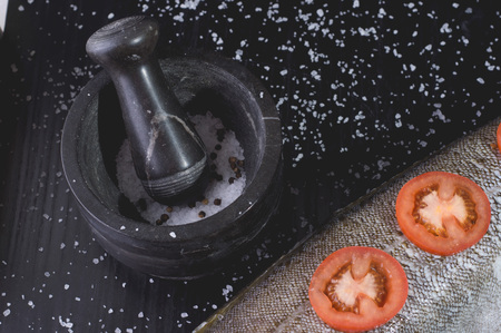 White fish with tomatoes and marble mortar on a black wooden background. Process of cooking